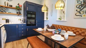 Une cuisine moderne avec des meubles de couleur bleu foncé, un four encastré et une table à manger en bois accompagnée d’un banc.