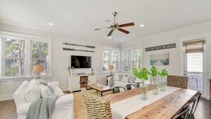 A spacious living room with a wooden dining table, white sofas, and a TV mounted on the wall.