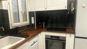 A kitchen with white cabinets, a black backsplash, a wooden countertop, a dishwasher, an oven, and a window with blinds. - Pleasant stay in the heart of Paris, 4 people max (Paris)
