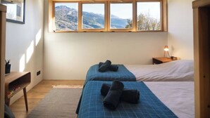 Un dormitorio con dos camas, un escritorio, una ventana con vistas a la montaña y una lámpara de noche.
