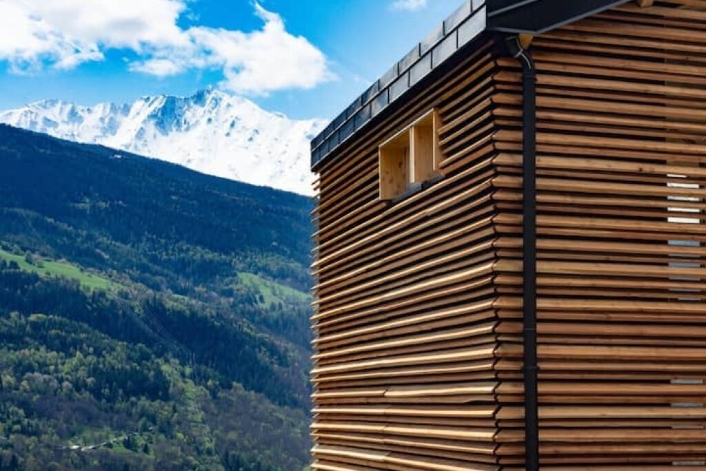 A wooden cabin with horizontal slats, a window, and a view of snow-capped mountains.
