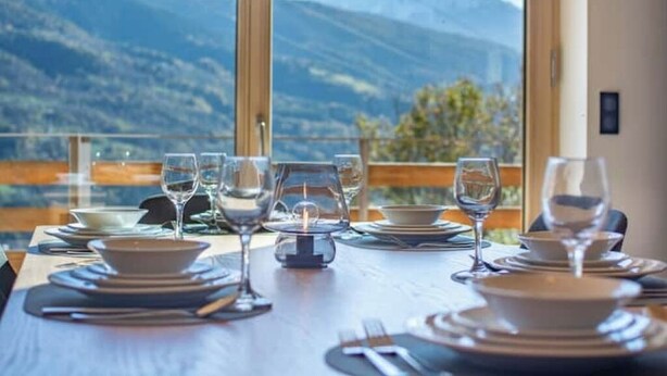 Mesa de comedor dispuesta con platos, vasos y cubiertos, con vistas a un paisaje montañoso a través de amplias ventanas.