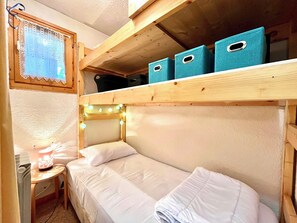 Uma cama beliche com cabeceira, uma pequena mesa de cabeceira com abajur e caixas organizadoras em uma prateleira de madeira acima da cama.