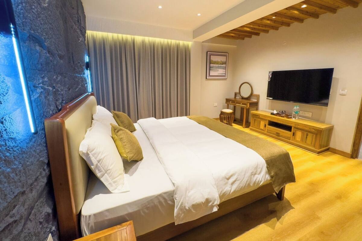A modern hotel room with a large bed, a wooden headboard, a flat-screen TV, a vanity with a mirror, and a large window with curtains.