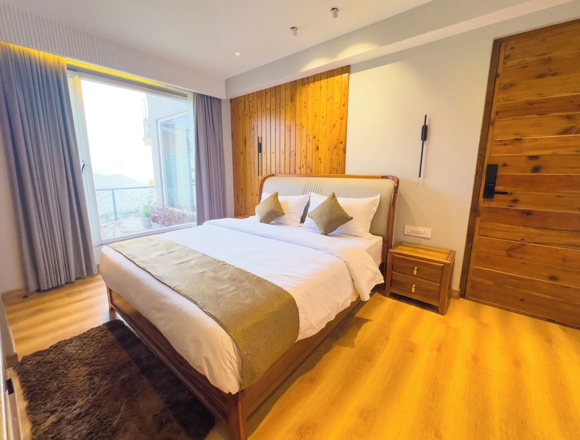 A modern bedroom with a large bed, wooden panel walls, a wooden wardrobe, and a view of the outdoors.
