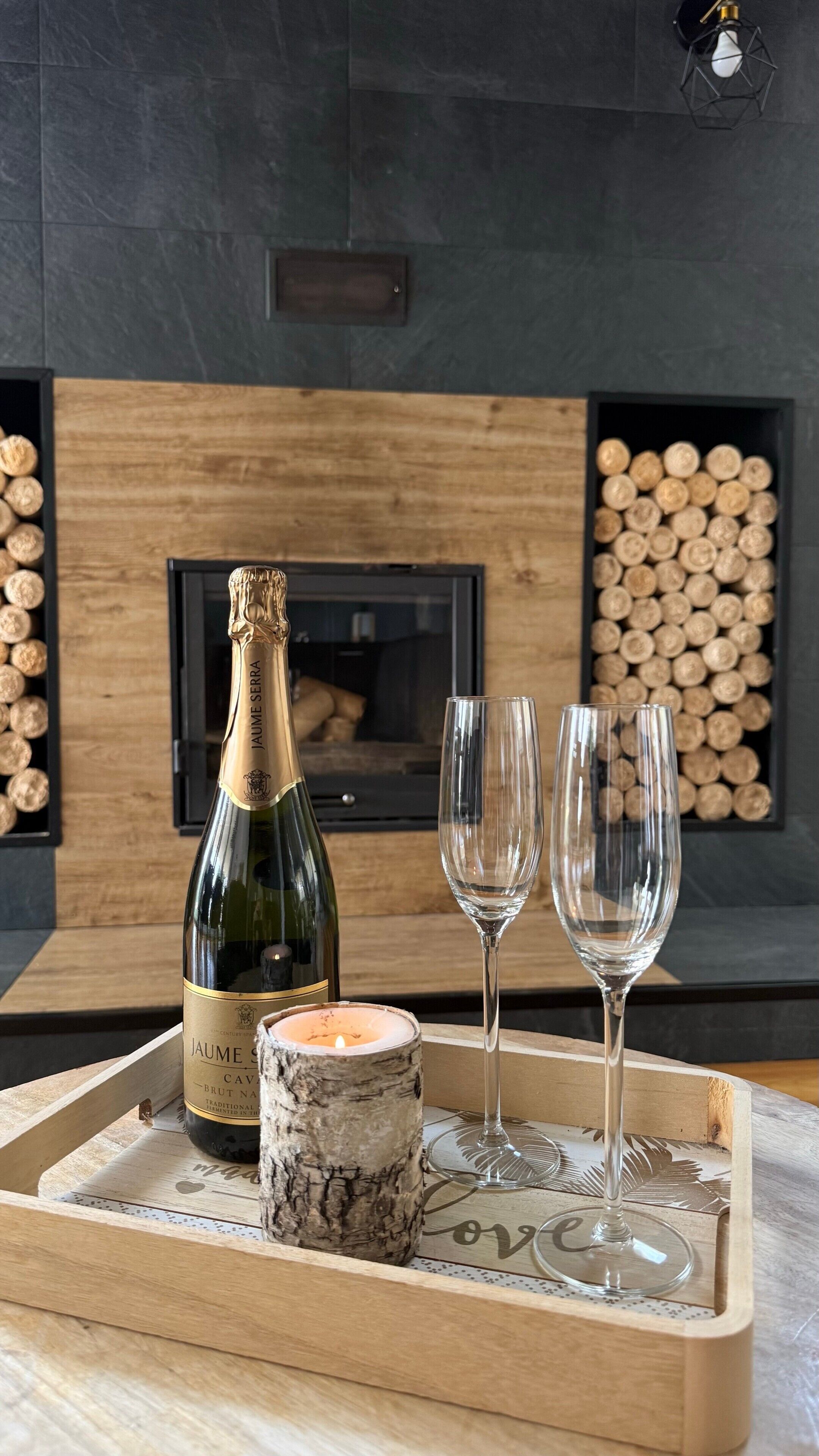 En træbakke med champagne, to glas og et lys på et bord foran en pejs.