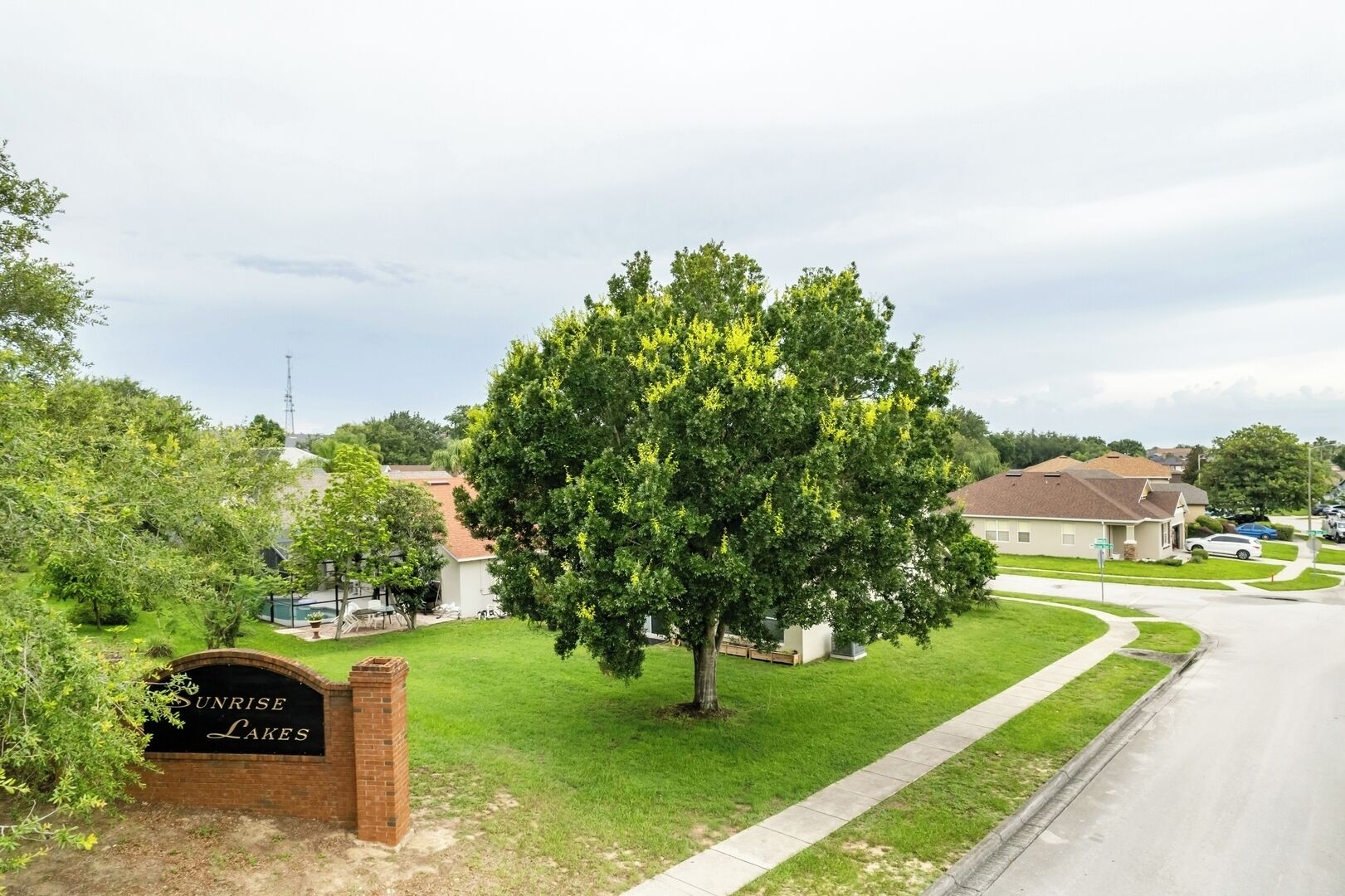 A residential area with a large tree in the foreground, a sign reading "Sunrise Lakes", and houses in the background.