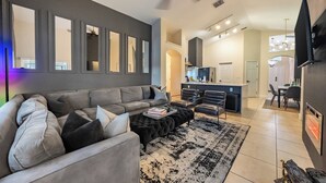 A modern living room with a grey sectional sofa, black ottoman, and a black and white rug. There is a dining area with a table and chairs, a kitchen with a bar, and a TV mounted on the wall.