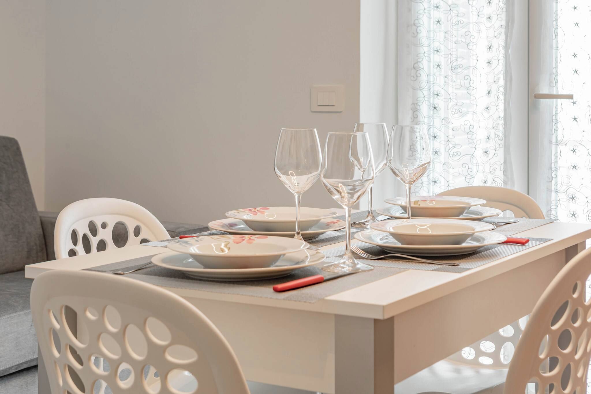 Una mesa de comedor dispuesta con platos, vasos y cubiertos, con una ventana y cortinas al fondo.