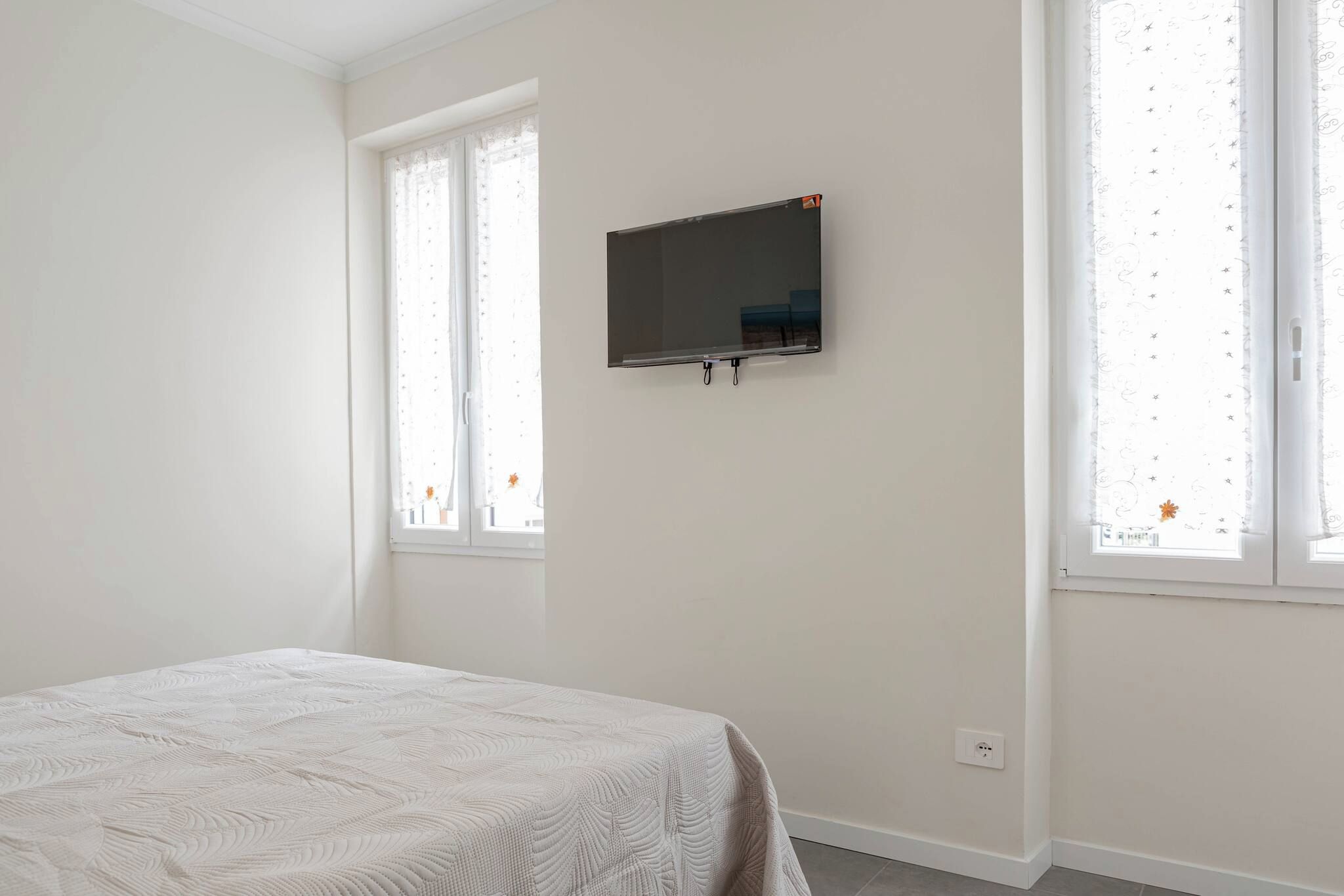 Une chambre à coucher avec un lit, un téléviseur fixé au mur et deux fenêtres avec des rideaux.