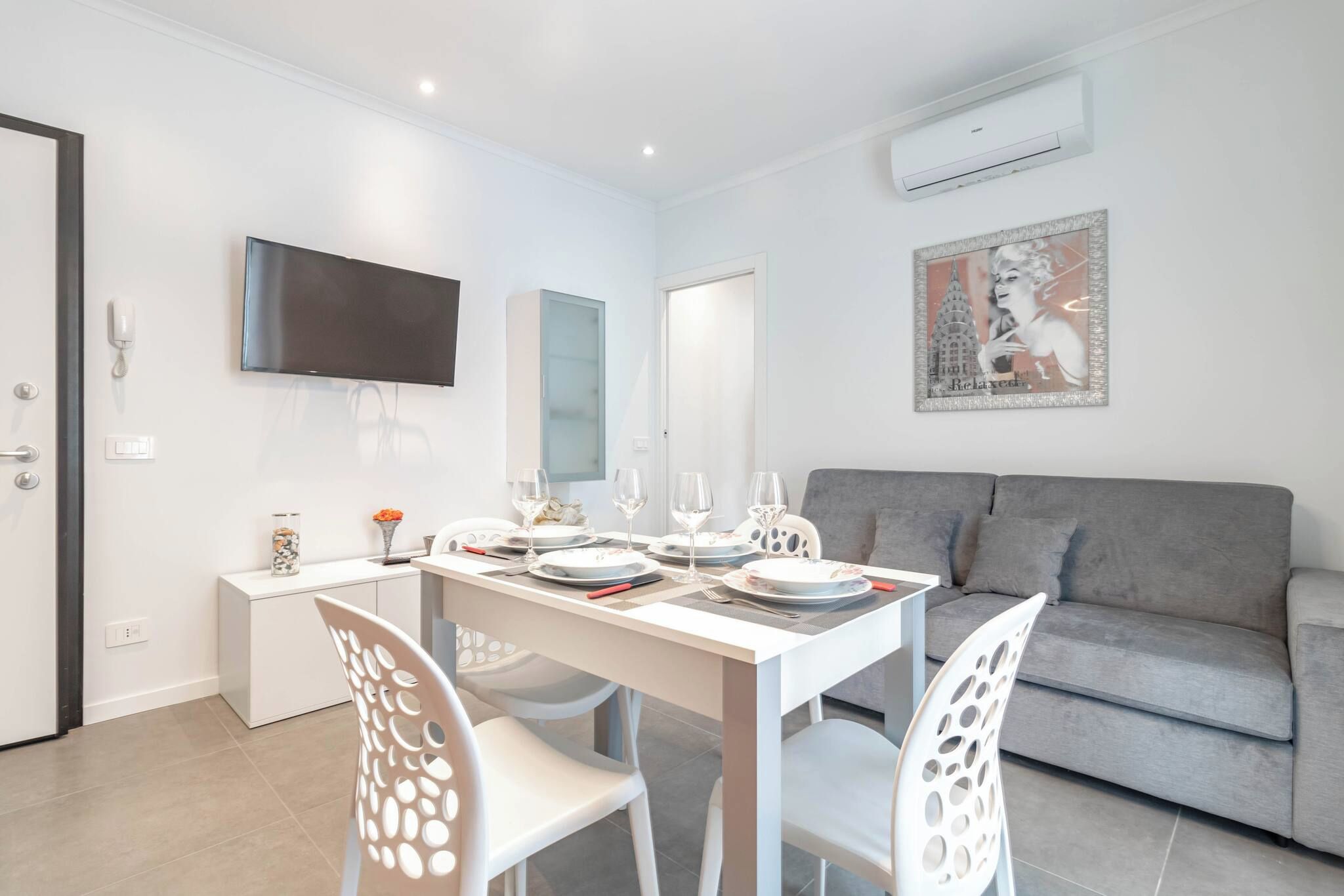 A modern living room with a grey sofa, a dining table set for four, and a flat-screen TV mounted on the wall.