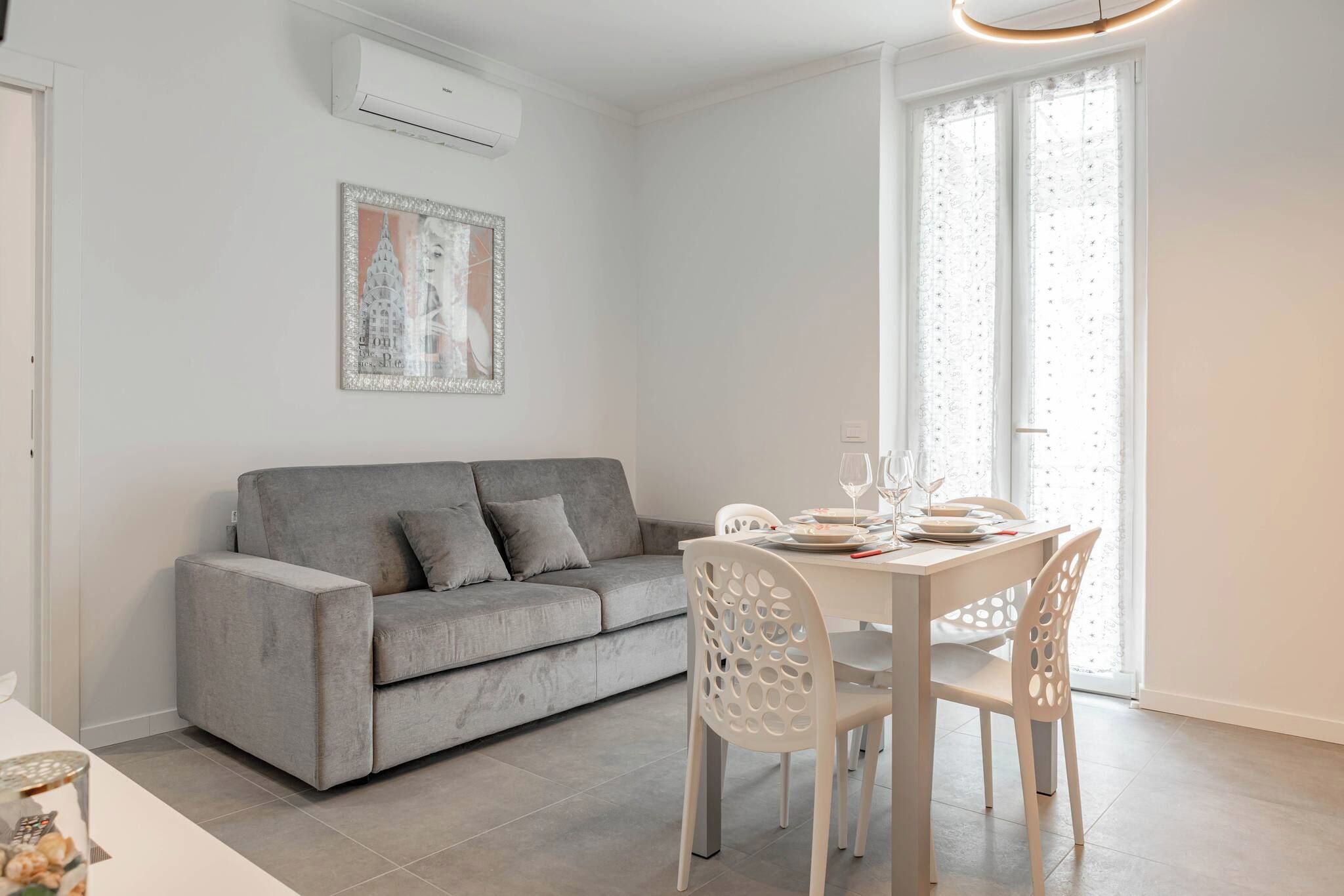 A modern living room with a grey sofa, a dining table set with white chairs, and a window with sheer curtains.