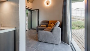 Un salon moderne avec un canapé gris, un luminaire mural rond et une vue sur l’extérieur grâce à une porte vitrée.