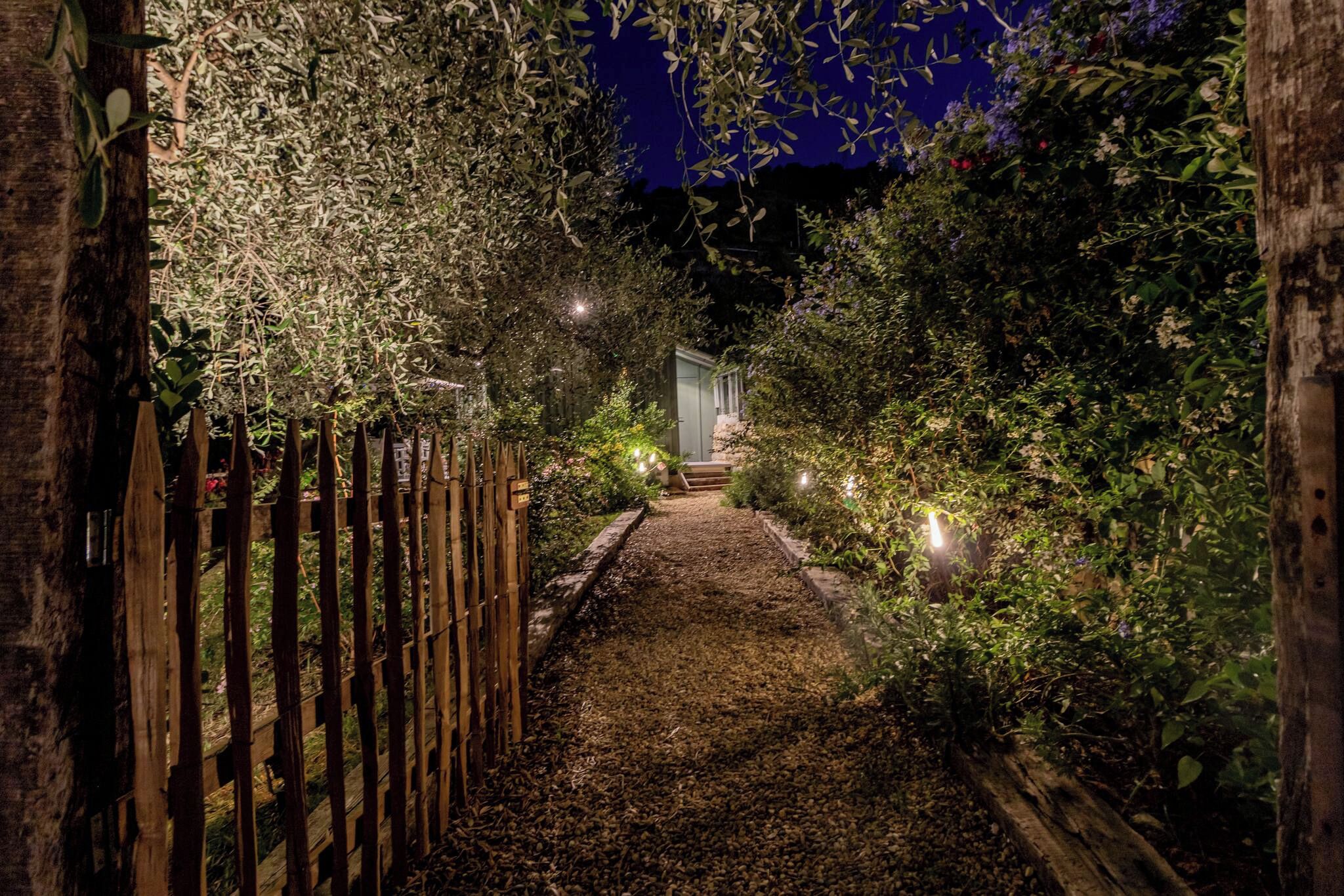 Un chemin illuminé serpentant à travers un jardin la nuit, menant à un bâtiment bordé d’une clôture en bois d’un côté et d’un feuillage dense de l’autre.