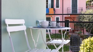 Ein Balkon mit weißen Stühlen und einem kleinen Tisch, mit Blick auf ein rosafarbenes Gebäude mit Balkonen und einem Garten.