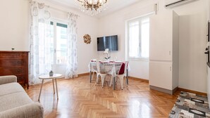 A spacious living room with a wooden floor, a dining table with chairs, a sofa, a chandelier, a TV, and an air conditioning unit. - Route 66 - Sea and Beaches (Sanremo)