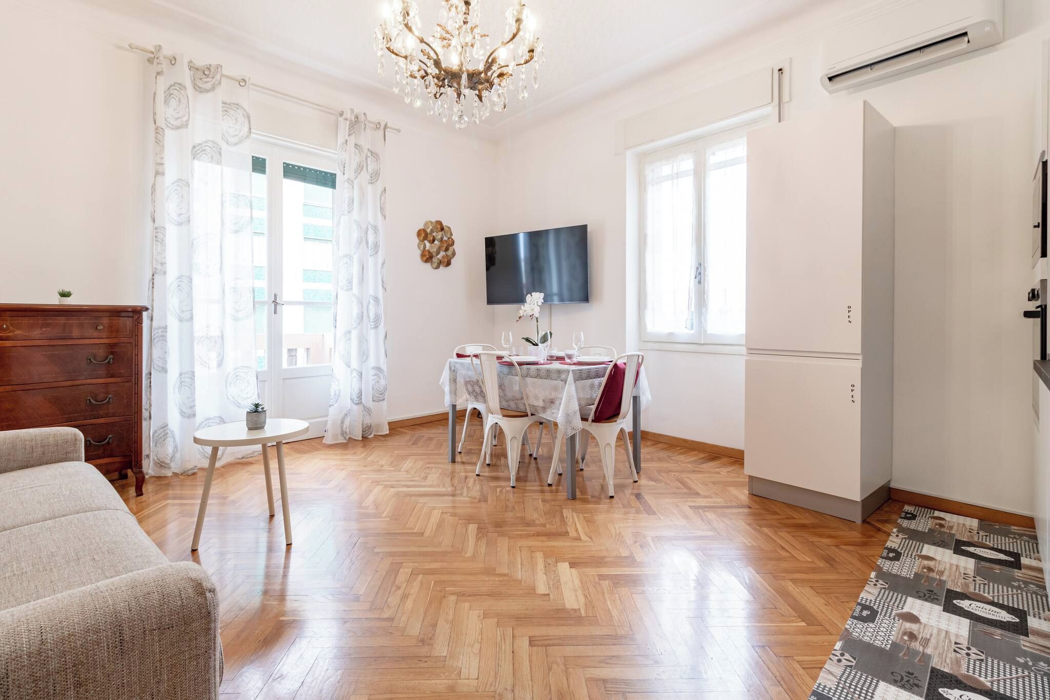 Une pièce avec une table à manger dressée pour quatre personnes, un lustre, un téléviseur fixé au mur et un plancher en bois.