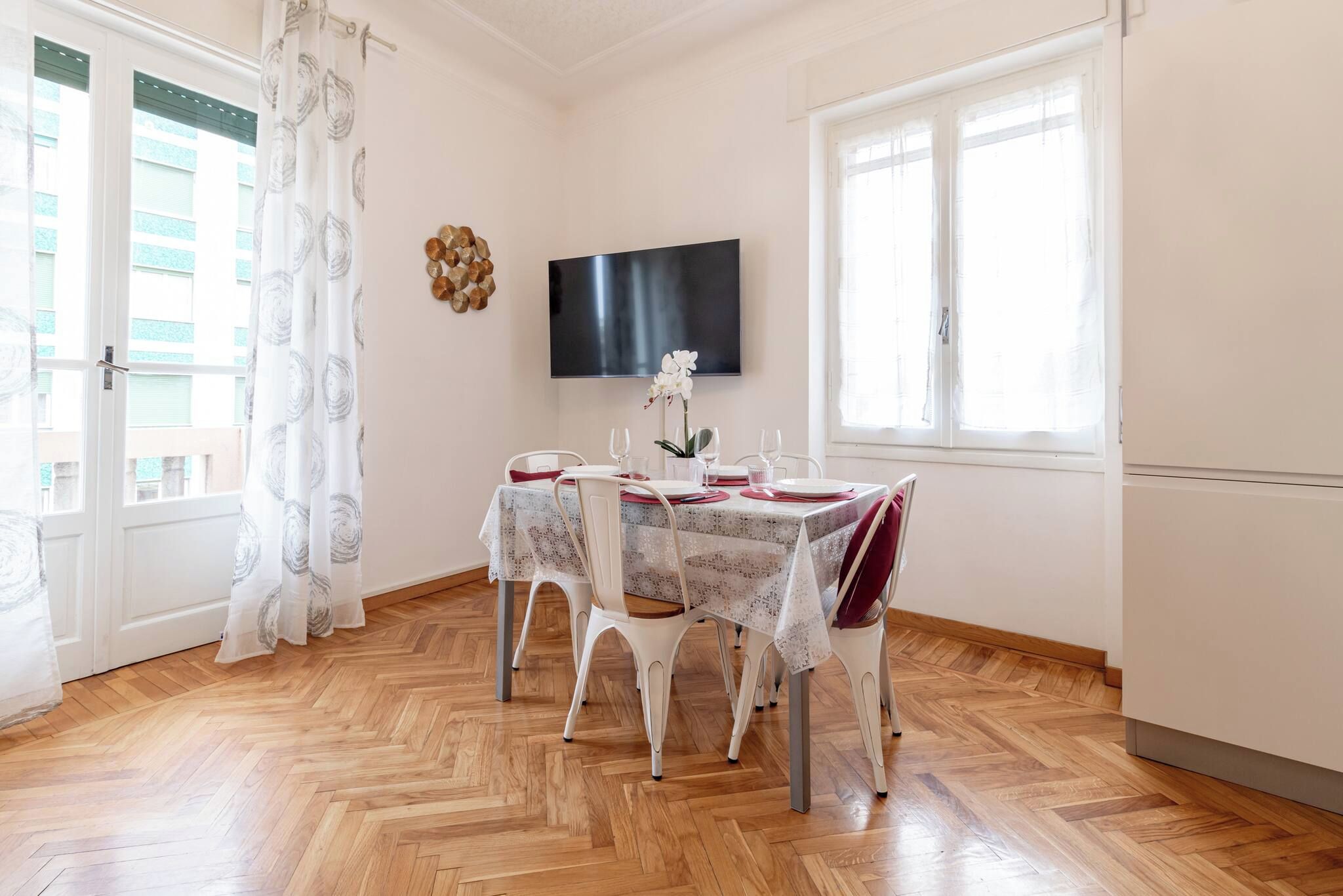A dining area with a table set for four, a flat-screen TV on the wall, and a window with curtains.