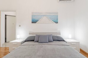 A bedroom with a bed, two bedside tables, a nightstand, a painting of a pier, and a door.