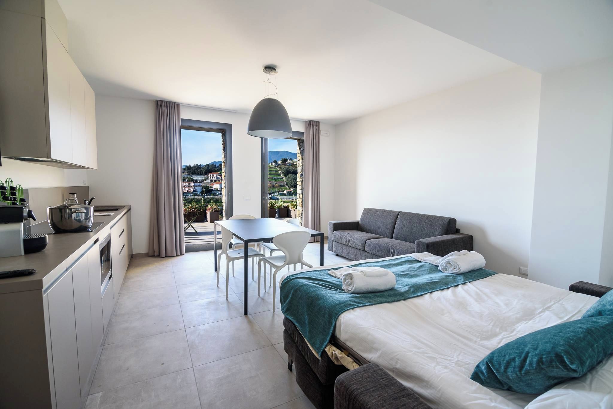 A modern hotel room with a bed, a sofa, a dining table, and a kitchen area.