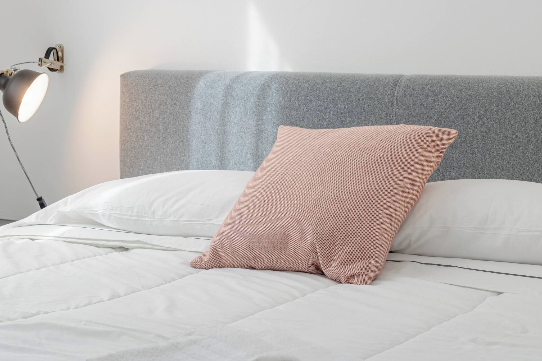 Un lit avec une tête de lit grise, une literie blanche et un seul coussin à motifs.