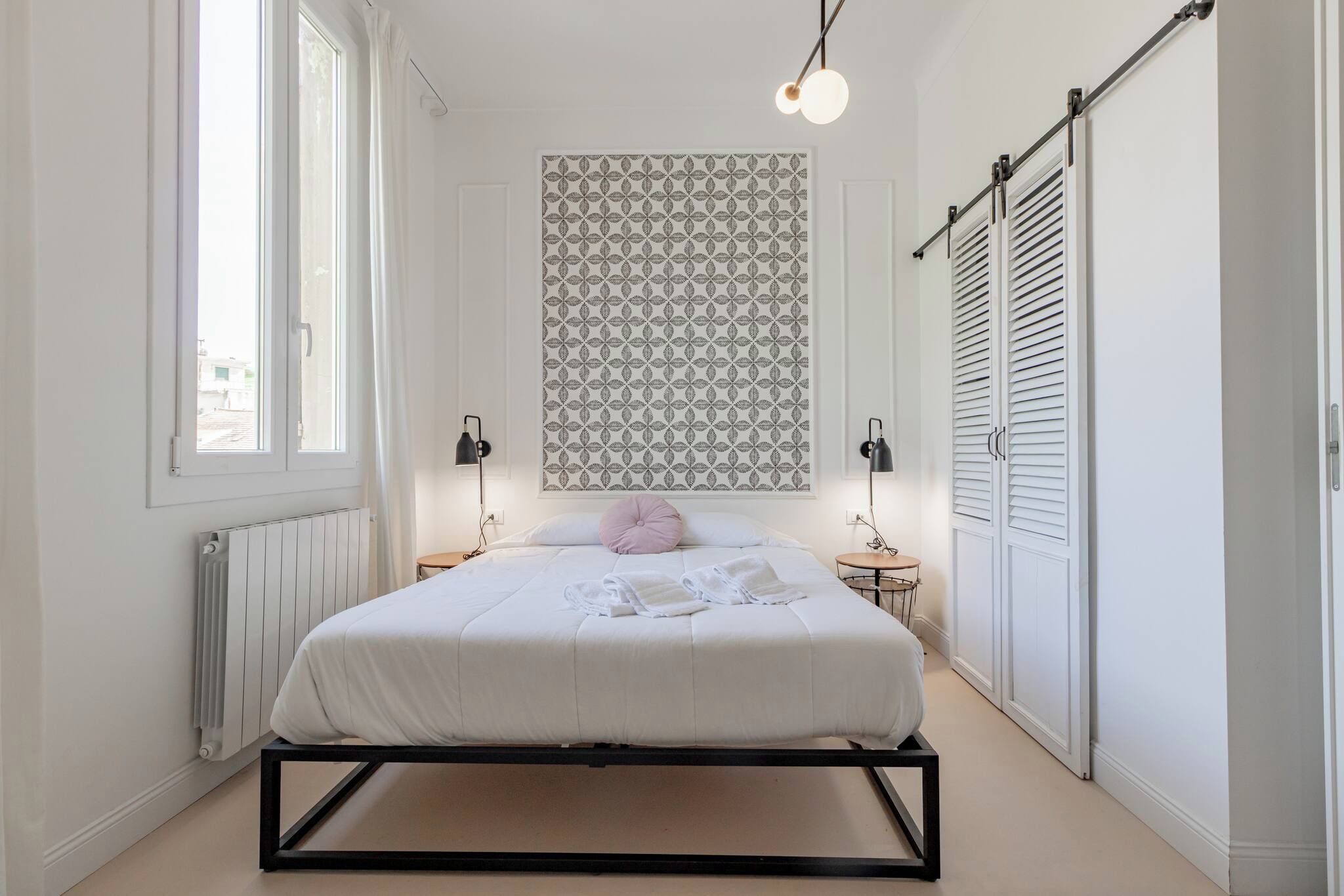 A bedroom with a bed, bedside lamps, a radiator, a window, and a patterned wall.