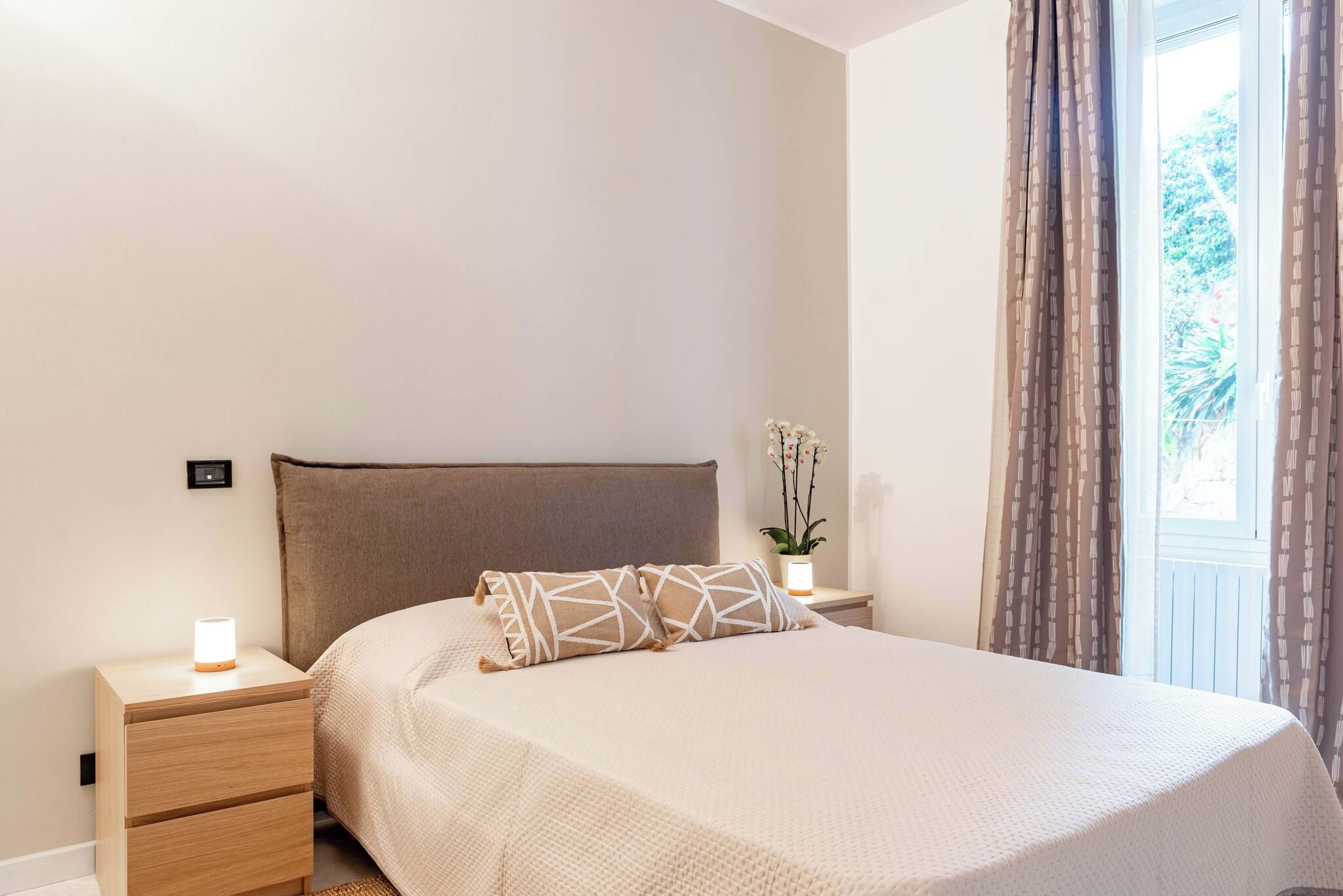 A bedroom with a bed, bedside tables, a nightstand, a lamp, and a window with curtains.