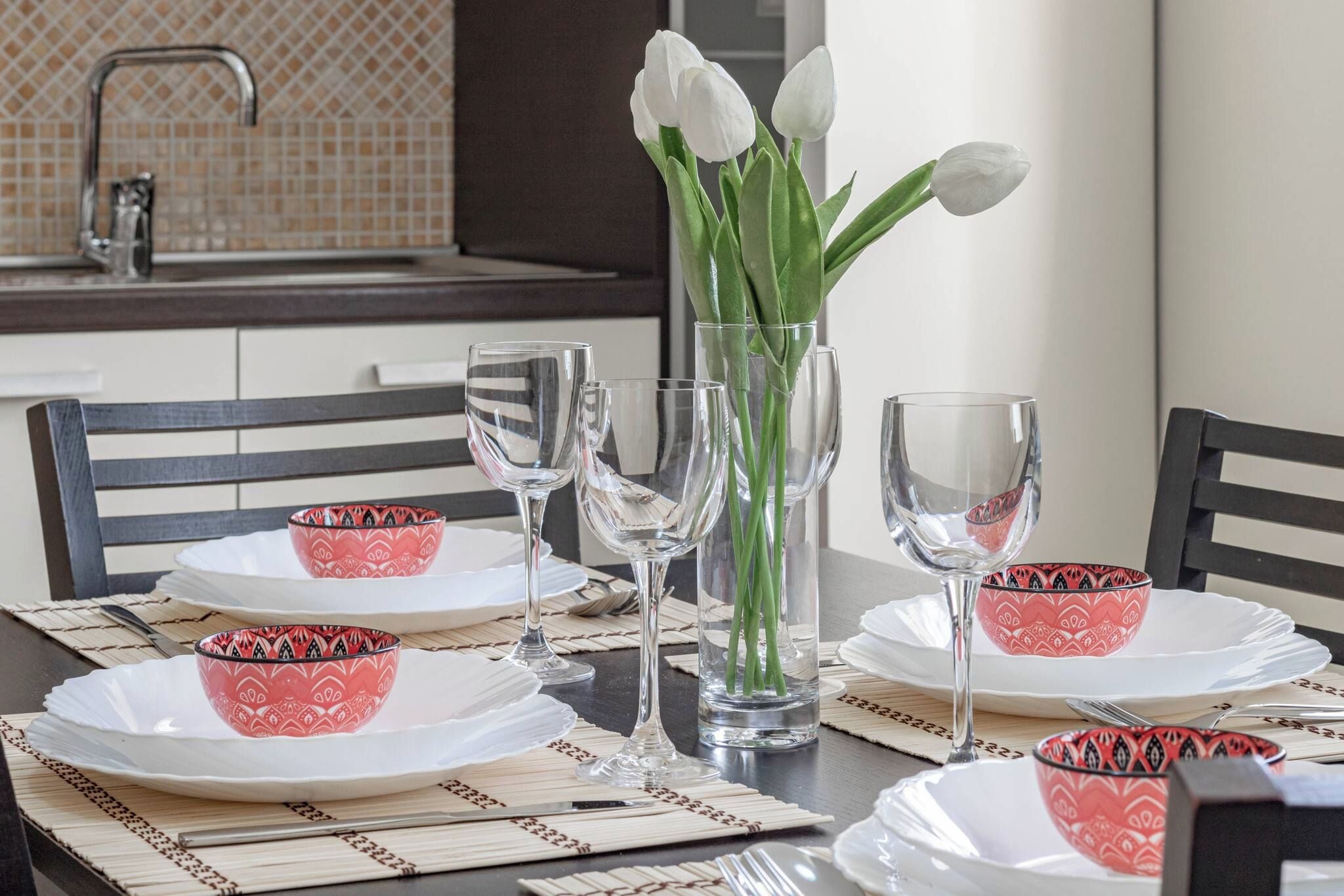 A dining table set with white plates, red patterned bowls, clear wine glasses, and a vase with white tulips.
