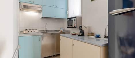 A compact kitchen with light blue cabinets, a stainless steel sink, a microwave, a kettle, and a refrigerator.