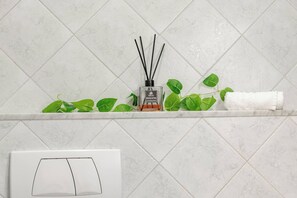 A bathroom with a white tiled wall, a shelf with a bottle of reed diffuser, green plants, and a white towel. - IL NIDO - Centro - Ariston (Sanremo)