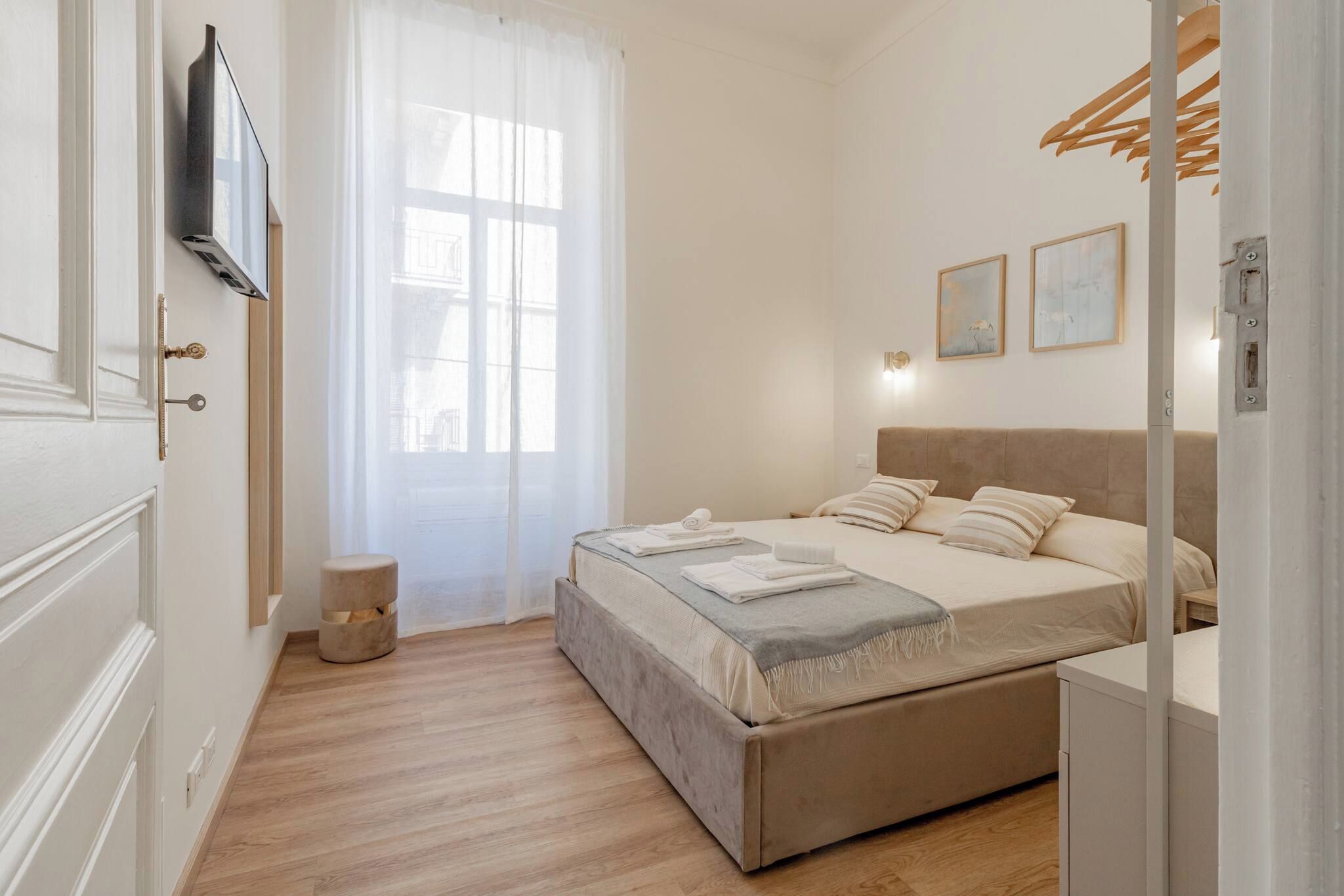 A bedroom with a bed, a bedside table, a wall-mounted TV, a window with curtains, and a small round stool.