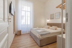 A bedroom with a bed, a bedside table, a wall-mounted TV, a window with curtains, and a small round stool.