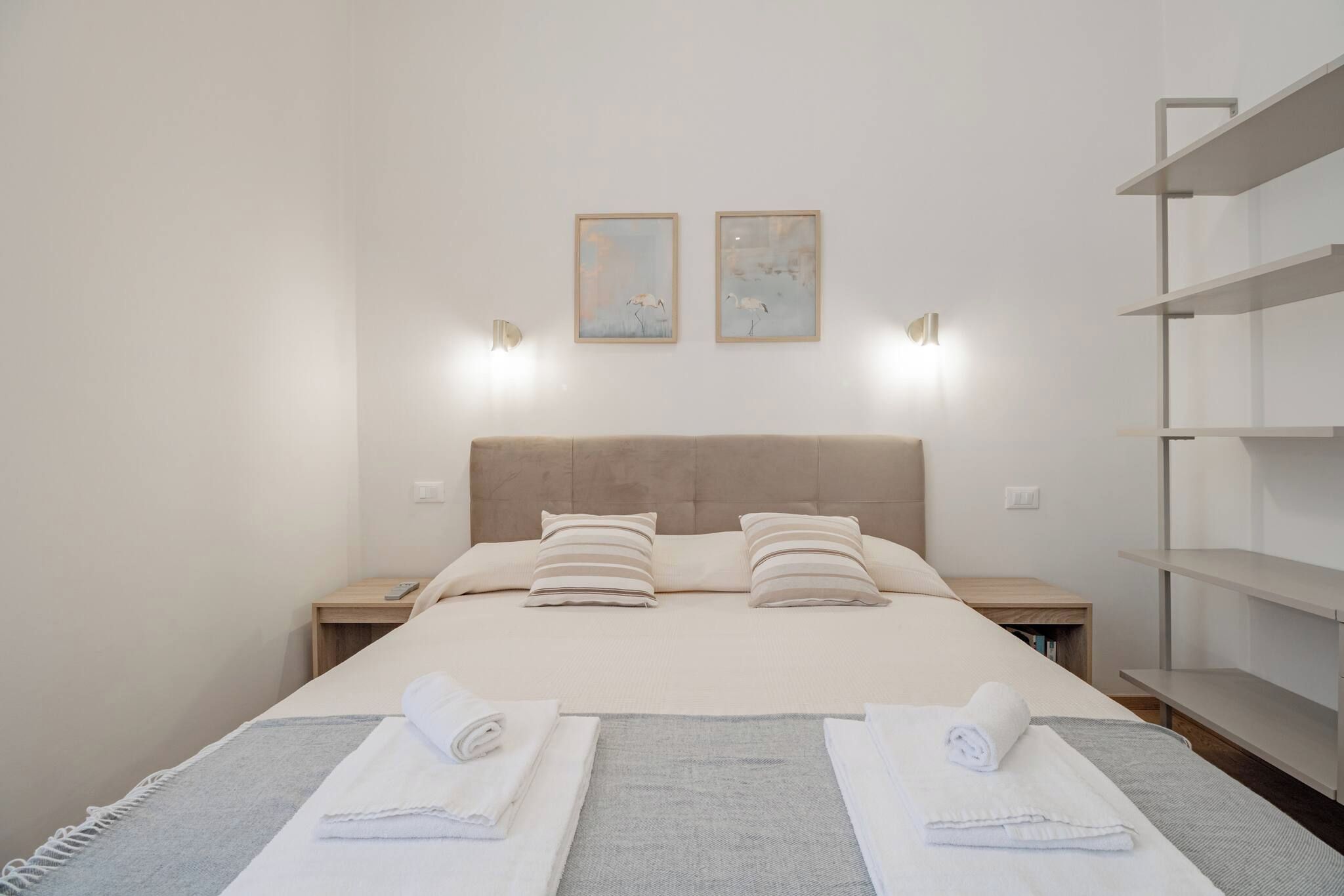 A hotel room with a bed, two bedside tables, a nightstand, a wall-mounted shelf, and two wall-mounted light fixtures.