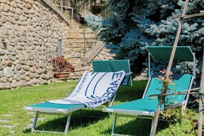 Two green lounge chairs with a towel draped over them, set against a stone wall and a coniferous tree.