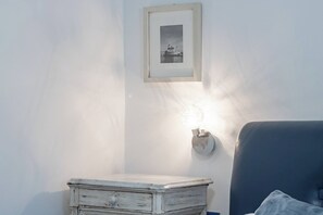 Quarto com mesa de cabeceira branca, quadro na parede e luminária fixada na parede.