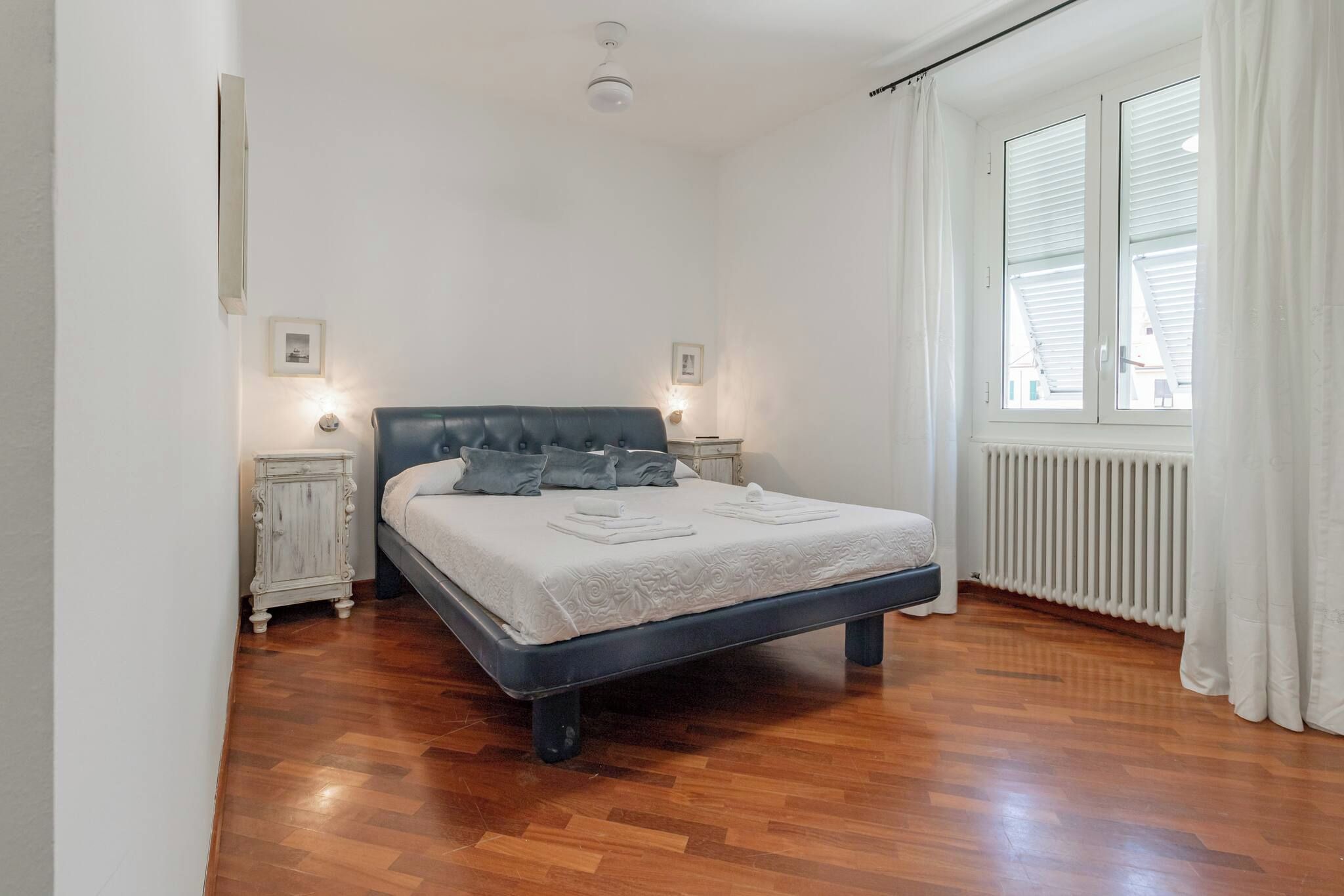 Un dormitorio con una cama, dos mesitas de noche, un radiador y una ventana con cortinas.
