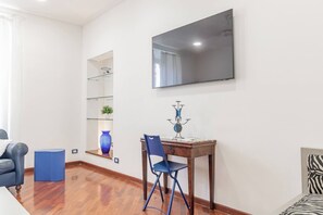 A room with a flat-screen TV mounted on the wall, a wooden desk with a chair, a blue vase on a shelf, and a zebra-patterned item on the floor.
