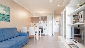 A modern living room with a blue sofa, a dining area with white chairs, a kitchen with white cabinets, and a TV on a white stand. - Silver House in Sanremo (Sanremo)