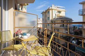 A balcony with yellow chairs, a small table, and a view of buildings and the sea.