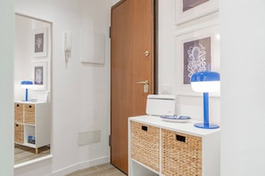 Un couloir avec une porte en bois, un mur blanc, une œuvre d’art encadrée, un meuble blanc avec des paniers et une lampe bleue.