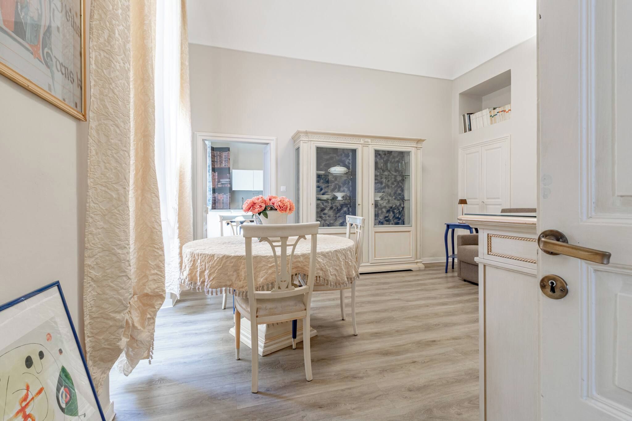 A dining area with a round table, chairs, and a vase of flowers. There is a cabinet with glass doors and a door with a brass handle.