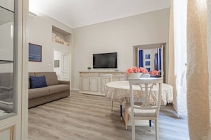 A modern living room with a sofa, a dining table with chairs, a television, and a view of a bedroom with a bed and curtains. - THE KINGDOM'S APARTMENT - Centro - Sanremo (Sanremo)