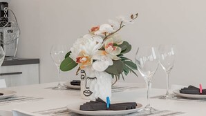 Ein Esstisch, gedeckt mit weißen Platzsets, einer Vase mit weißen Blumen und leeren Weingläsern.