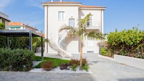 Ein modernes, zweistöckiges Haus mit weißer Fassade, einer Wendeltreppe und einem Garten mit verschiedenen Pflanzen und einem Palmenbaum.