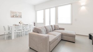 Ein modernes Wohnzimmer mit einem beigefarbenen Ecksofa, einem Esstisch mit Stühlen und großen Fenstern, die für natürliches Licht sorgen.