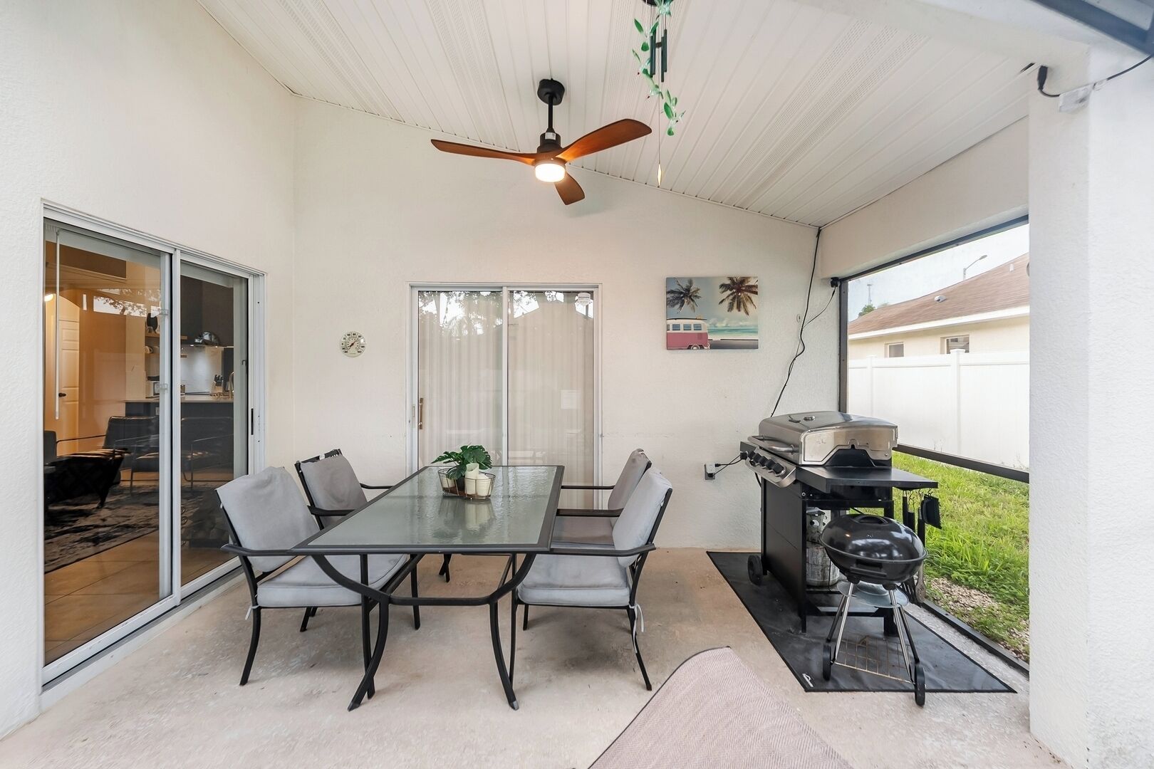Eine überdachte Terrasse mit Glastisch, vier Stühlen, Deckenventilator, Grill und einer Glastür, die zu einem Innenbereich führt.