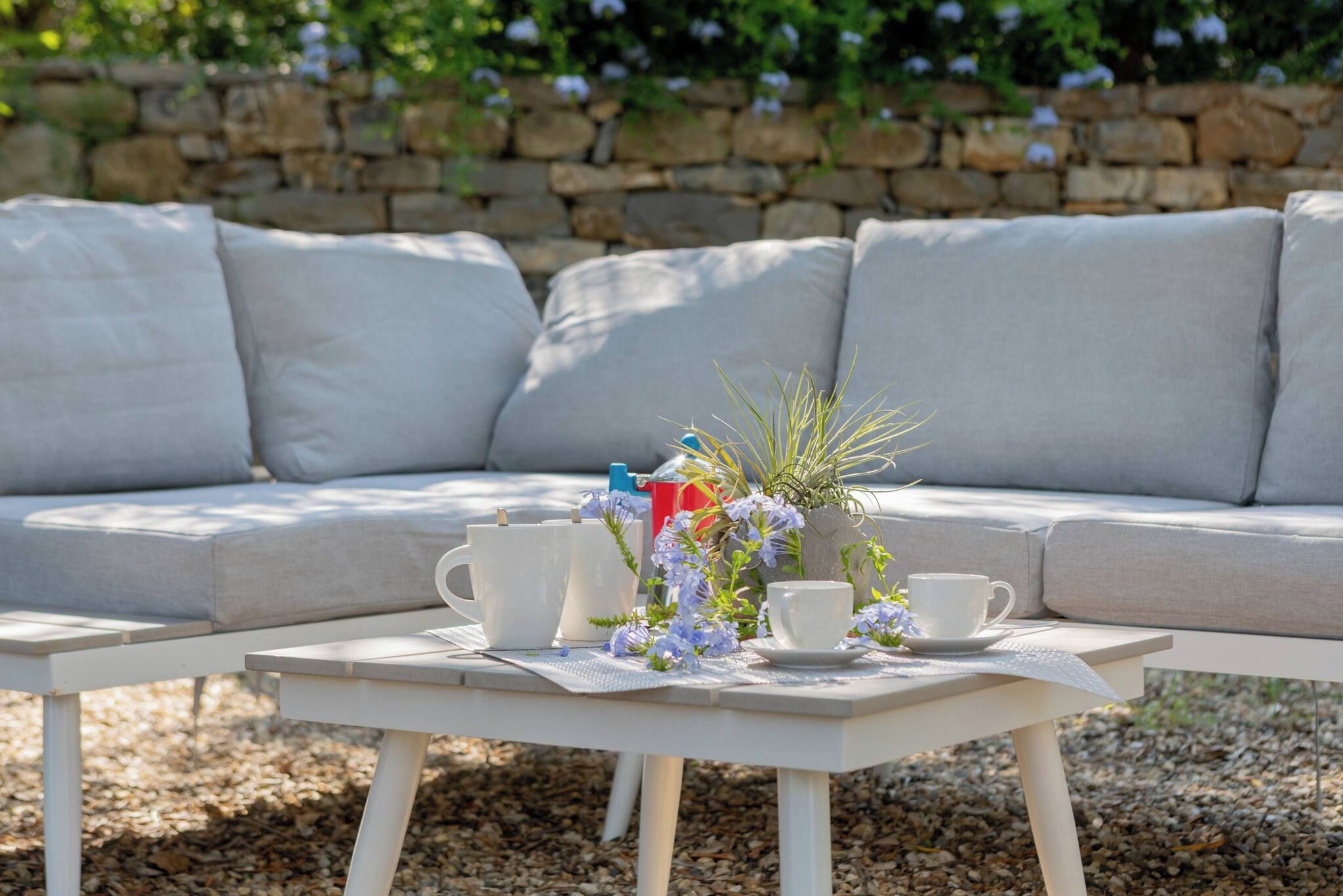 A white outdoor seating area with a table set for two, featuring a vase with flowers and a red mug.