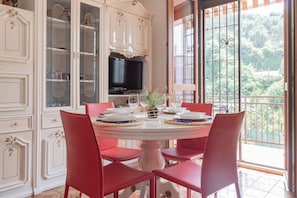 Un coin repas avec une table ronde, des chaises rouges, un téléviseur et une vue sur la verdure par une fenêtre.