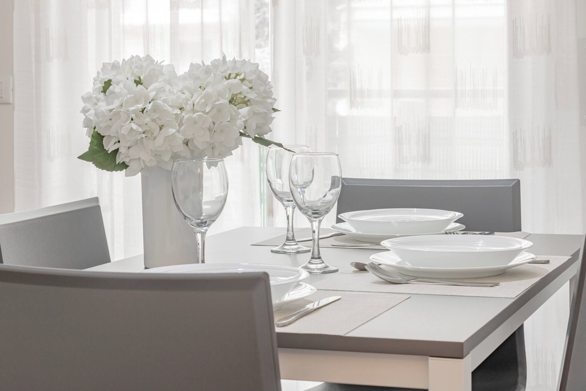 A dining table set with white plates, clear wine glasses, and a vase of white flowers.