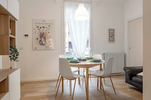 A dining area with a table set for four, surrounded by white chairs, a pendant light, and a window with sheer curtains.